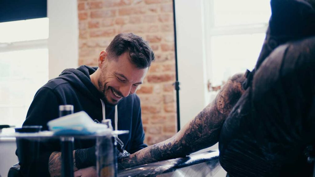 Smiling tattoo artist tattoos wrist of client. 