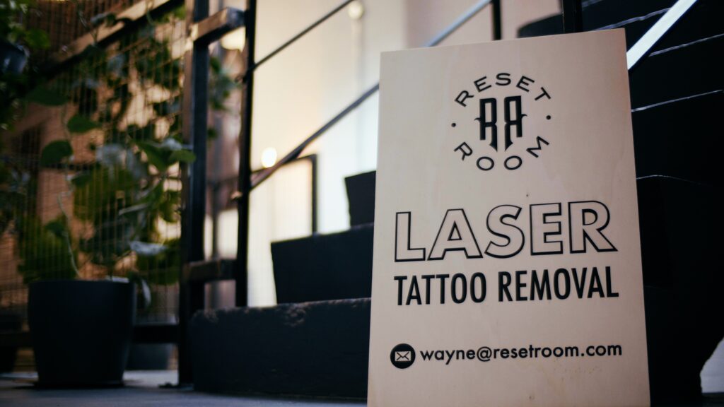 A wooden signboard with the text "RESET ROOM LASER TATTOO REMOVAL" and an email address for contact is placed near the entrance of a staircase. The background features plants and a metal railing, creating a welcoming atmosphere for those seeking laser tattoo removal.