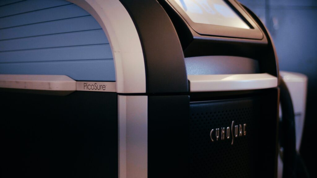Close-up of a PicoSure laser machine, primarily black with white and blue accents. The machine features a display screen on the top and the PicoSure logo prominently visible on the side. 