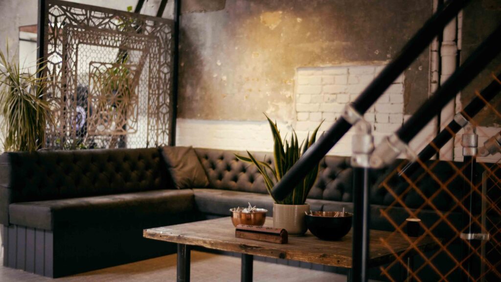 The waiting area of No Regrets Tattoo Studio London. Images shows a grey corner sofa, a wooden table with a plant and bowls of sweets in the middle of the table. In the foreground of the image is the banister of stairs. 
