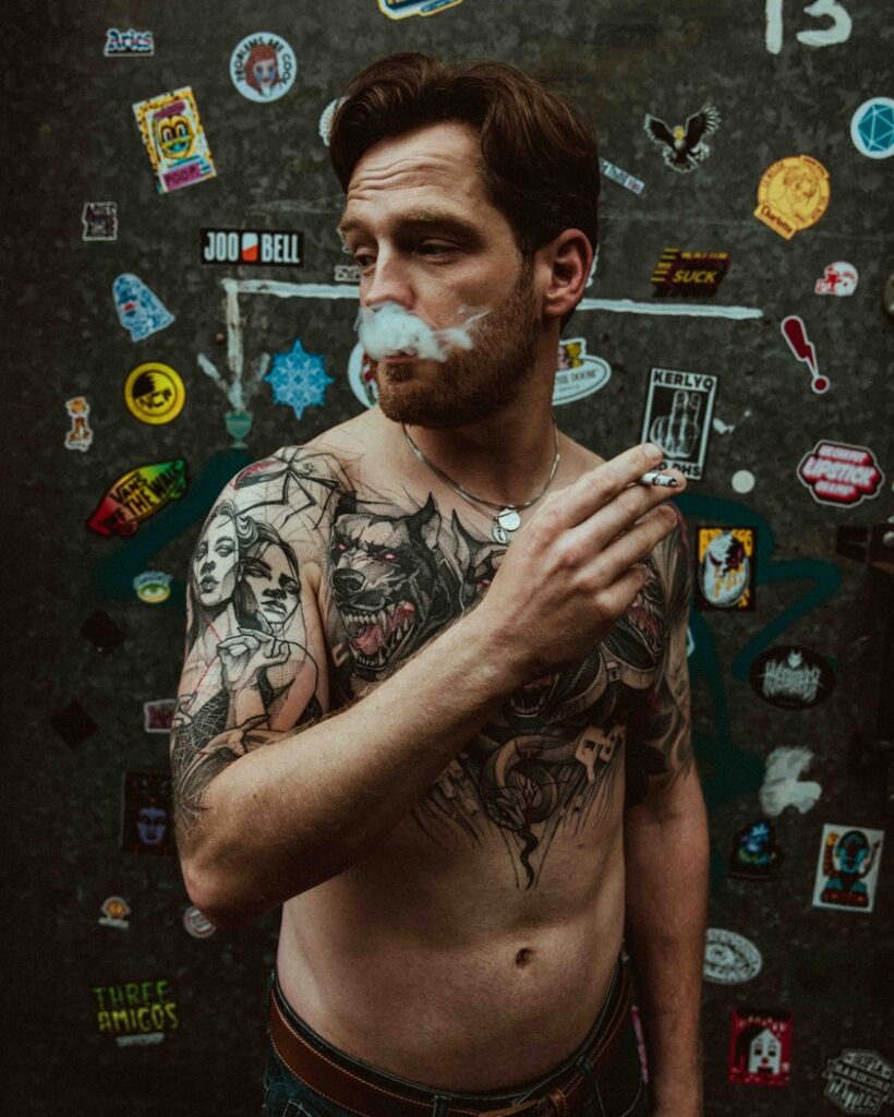 Jake stands in front of  a wall with stickers on, he looks to the side and has a cigarette in his hand. Smoke is coming out of his mouth. 
