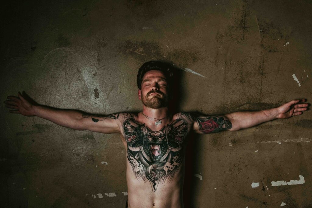 Jake lies on a stone floor with his arms spread out. His Greek Mythology tattoos can be seen on his chest. 
