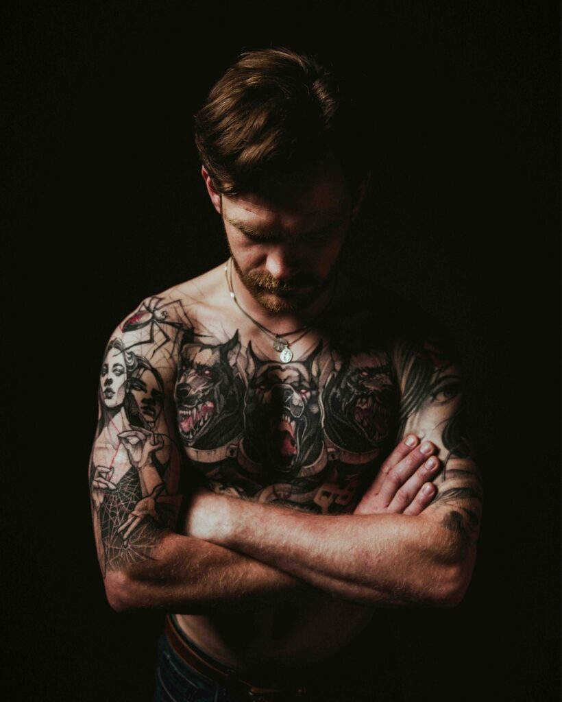 Jake stands against a black background. He is topless, looking down with his arms crossed. You can see his mythology tattoos on his chest and upper arms.