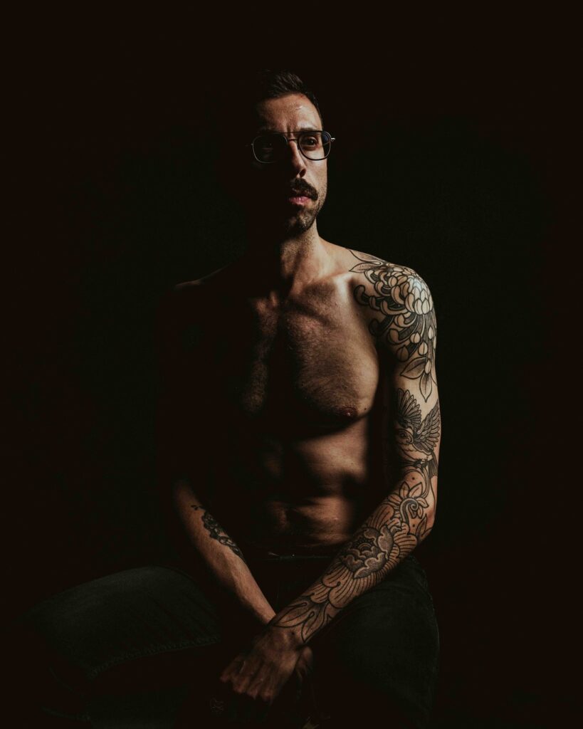 Luca sits in front of a black background on a stool. Half of his body is in the shadow, while you can clearly see the detail of his sleeve tattoo.