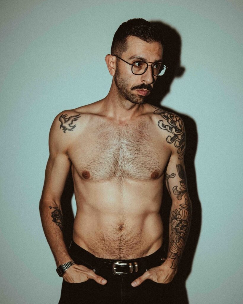 Luca stands against a white background face onto camera topless, looking to the left. On his left arm, you can see his black line floral sleeve by Giovanna.