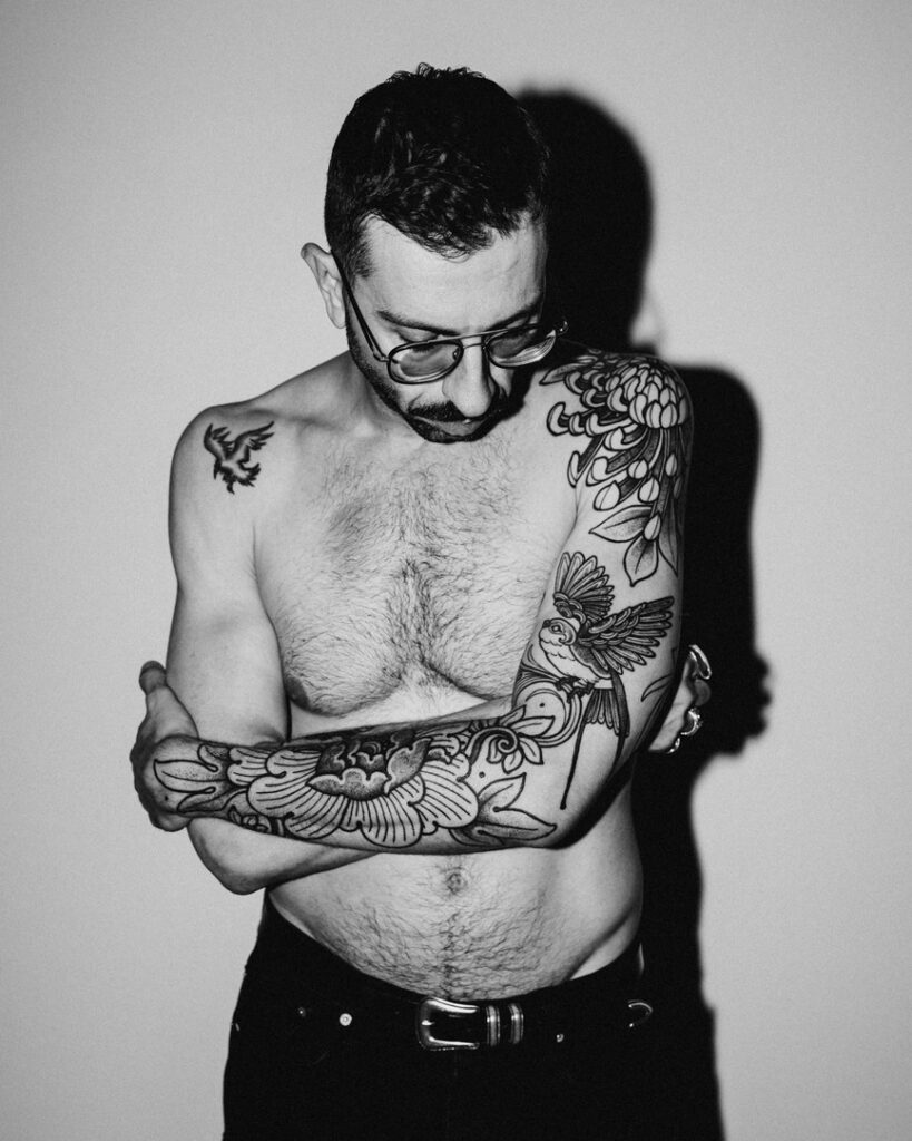 A black and white image of Luca standing with his arms folded while he looks down. You can see his black line floral sleeve on his left arm.
