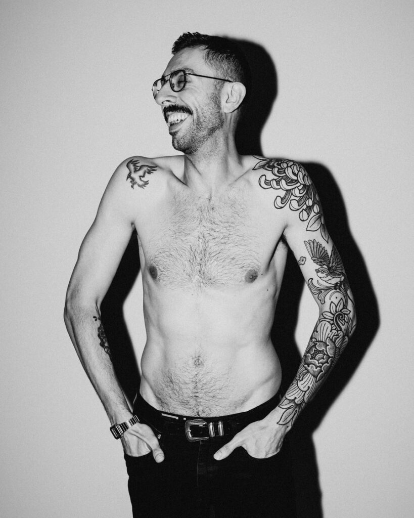 A black and white image of Luca with his hands in his pockets. He looks to the right laughing.