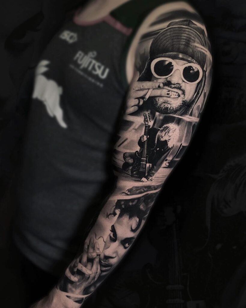 Music tattoos: Image of a man with a black and grey sleeve tattoo that shows Kurt Cobain wearing a hat and white round sunglasses and also him sitting on the ground with his guitar. At the bottom of the arm you can see a portrait of Prince