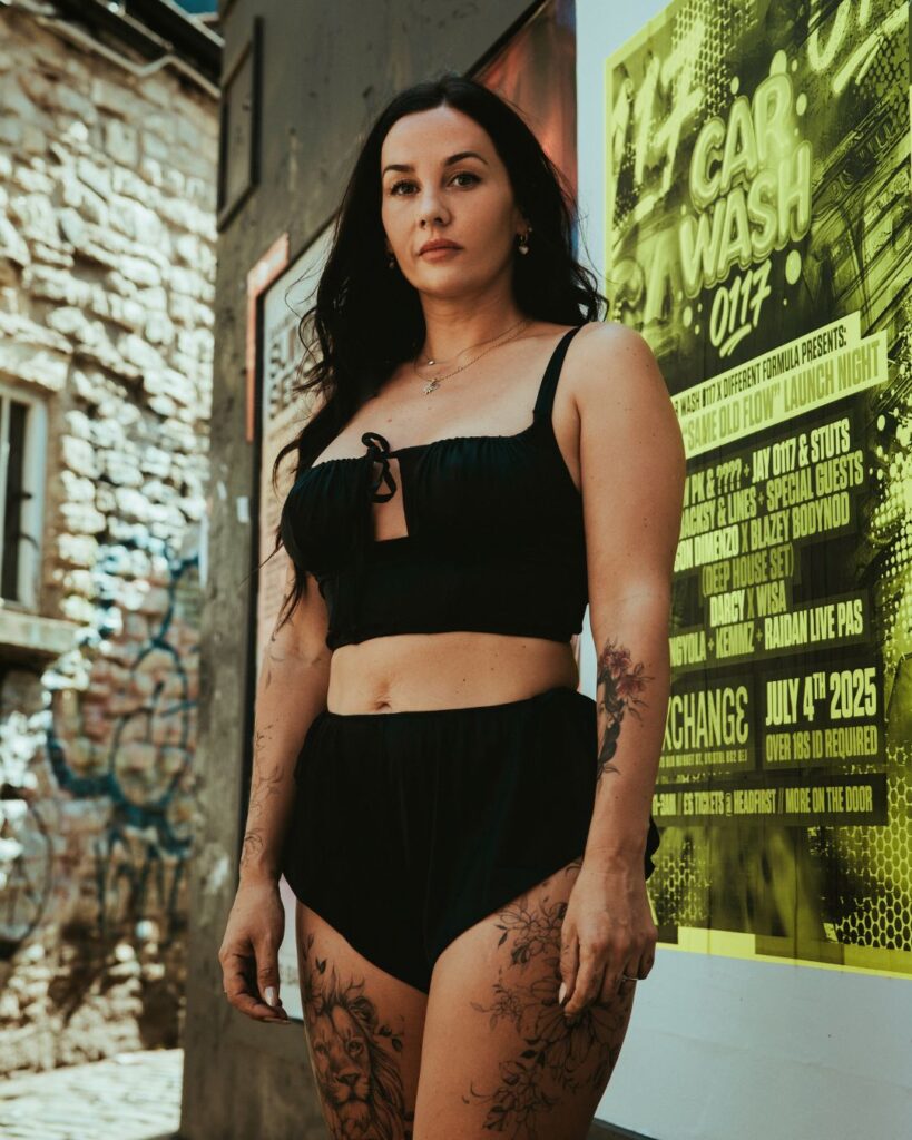 Stacey stands face on to the camera in a black crop top . Her floral and lion tattoo designs are visible on her thighs and arms. 