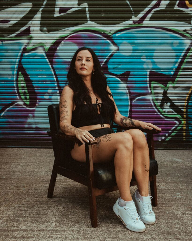 Stacey sits in a chair in front of a shutter covered in graffiti. Her fine line tattoos are shown on her right arm.