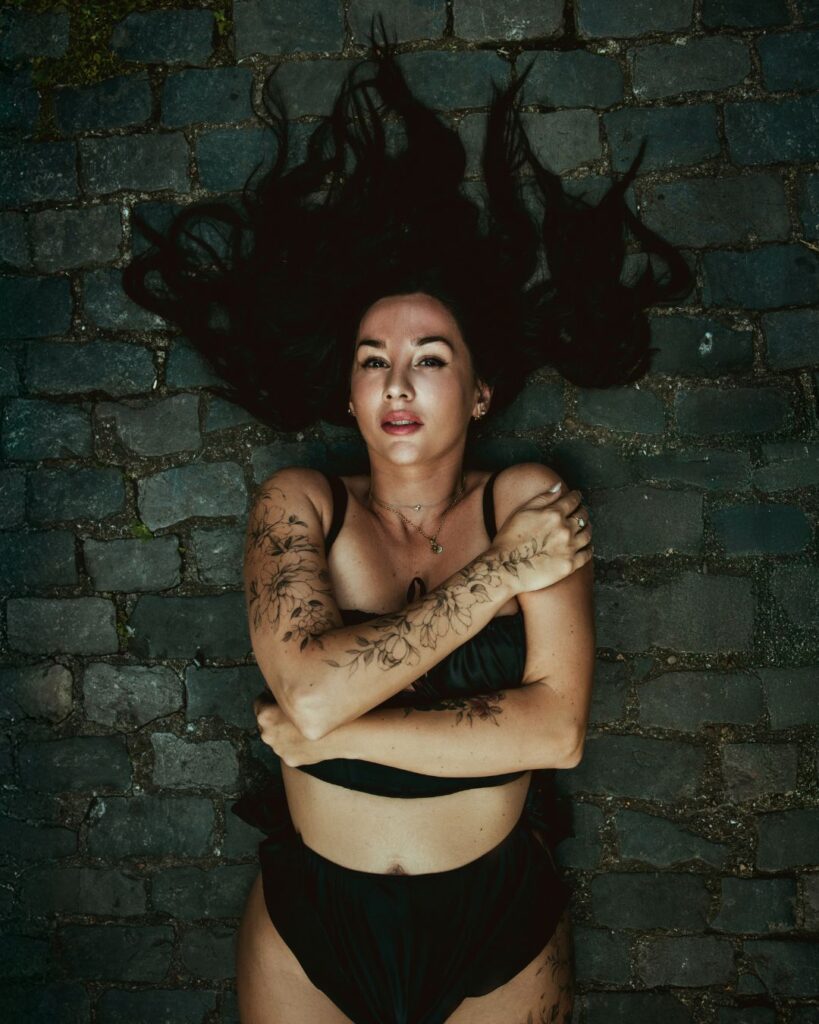 Stacey lies on a grey cobbled path with her arms wrapped around her chest. You can see her floral tattoo designs on her right arm.