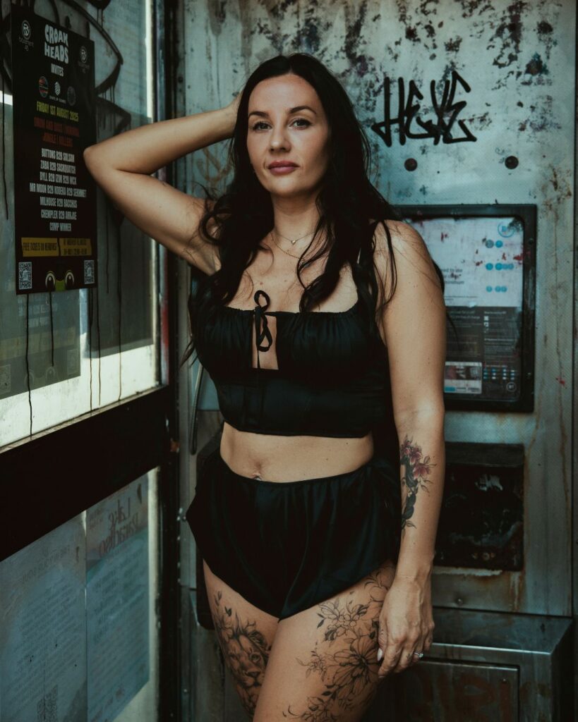 Stacey stands in a phone box with her arm raised above her head. You can see her fine line tattoos on both thighs and left forearm.