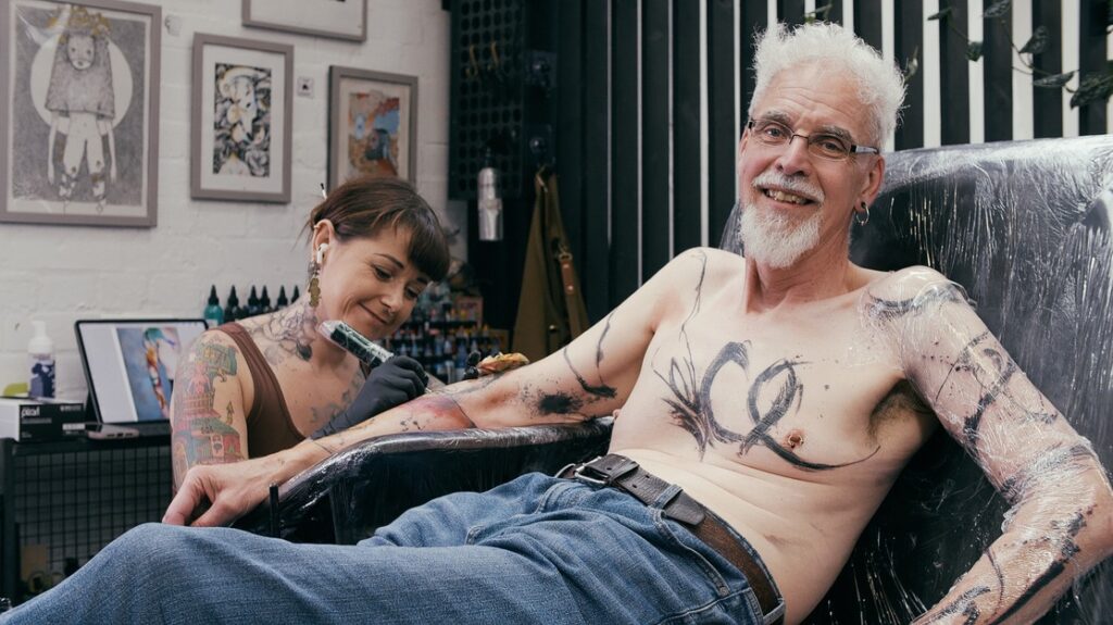 Steve sits up on a tattoo bed smiling at the camera while Tembo, a female tattoo artist tattoos his arm.