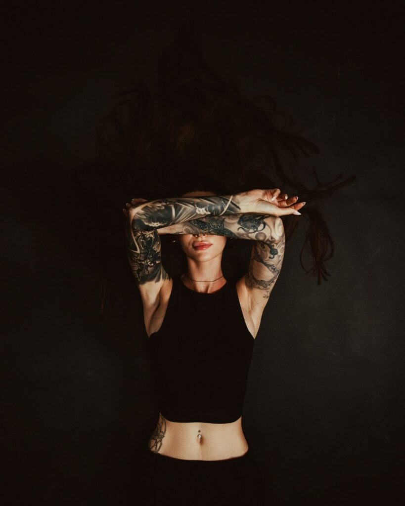 A woman lies on a black background with her tattooed arms crossed over her face so her eyes aren't viewable.