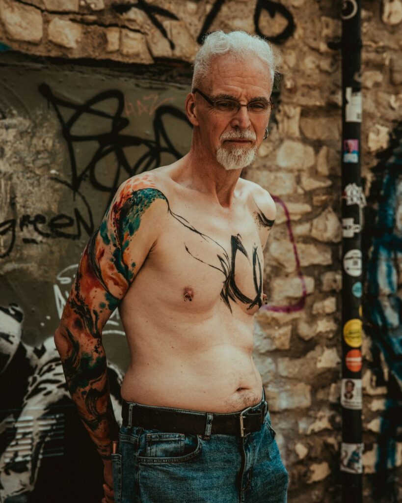 Steve stands against a brick wall, topless with his arms behind his back. His left arm shows his first tattoo, colourful abstract paint strokes.