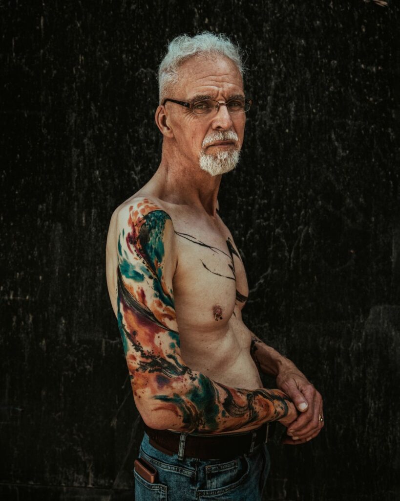 Steve stands side on against a black background. His right arm is on show and it shows his first tattoo which shows colourful abstract paint strokes.