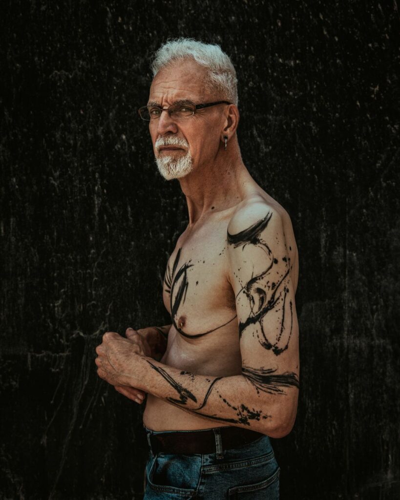 Steve stands side on against a black background. His left arm is on show and it shows his first tattoo which shows black paint strokes.