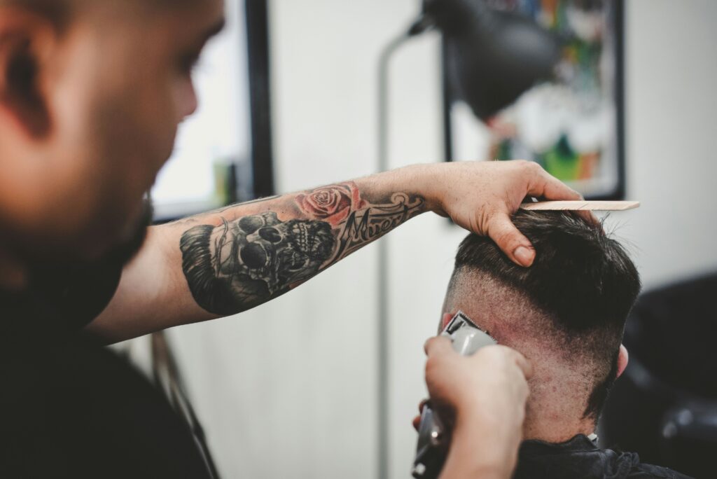 Barber with forearm skull tattoo holds head of male client while cutting his hair. 