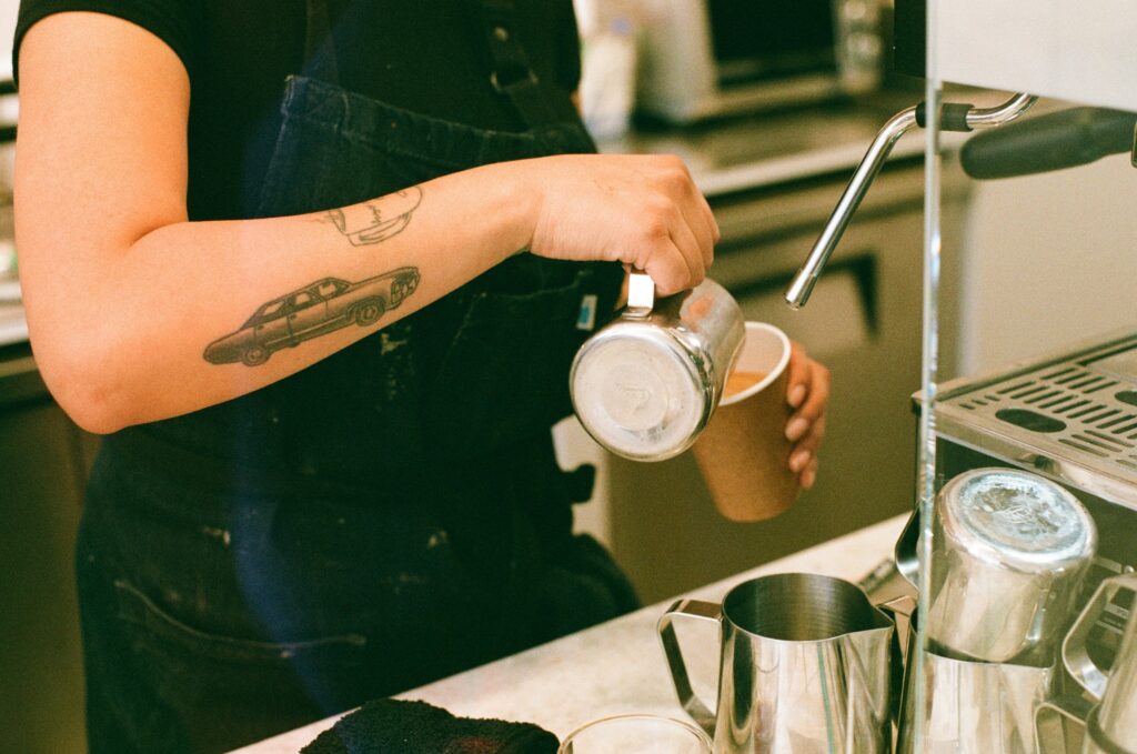 Barista with car tattoo on arm, pours milk into a takeaway coffee cup. 