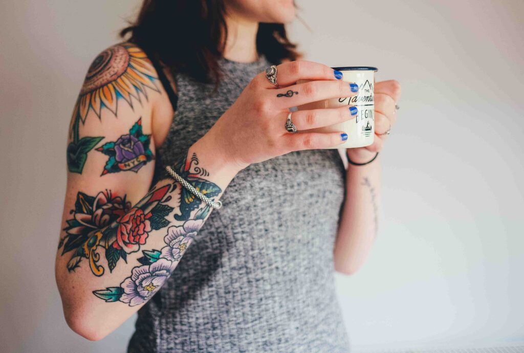 woman standing in front of a light background holding a mug. Wearing a grey tank top with neo traditional colourful flower tattoos on arm, 