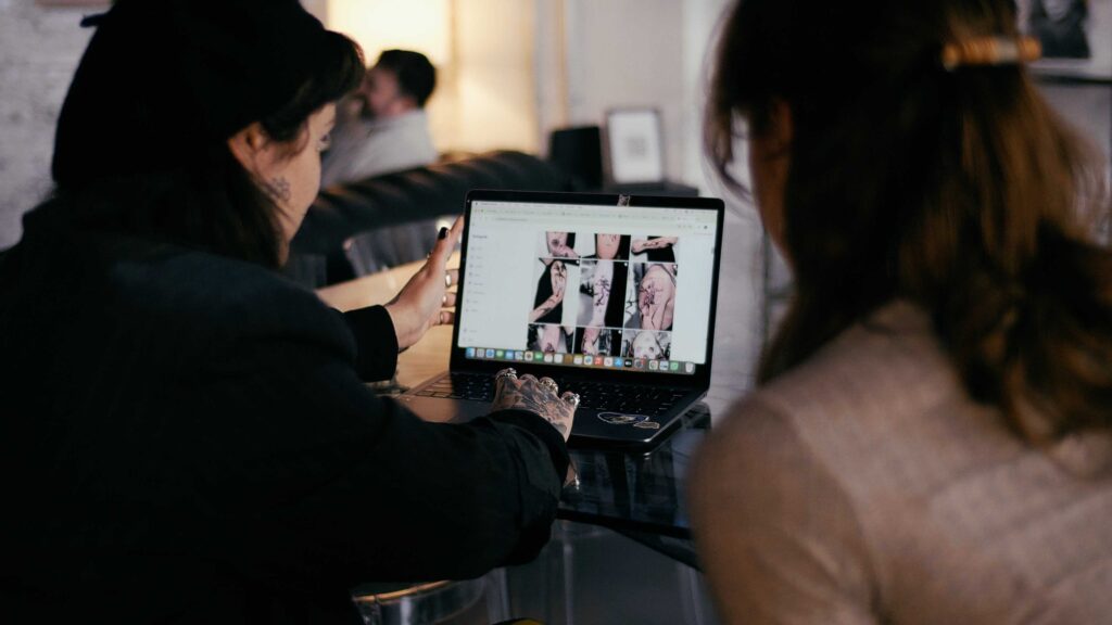 A female tattoo consultant and female client look at a tattoo artists work on a laptop for first tattoo ideas. 