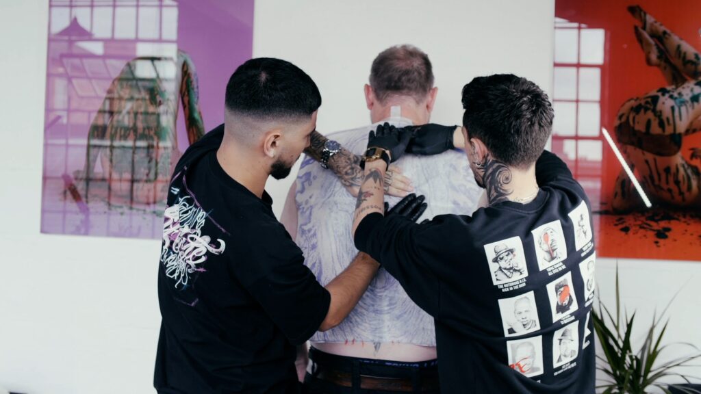 Two tattoo artists apply a large stencil to a client's back. 