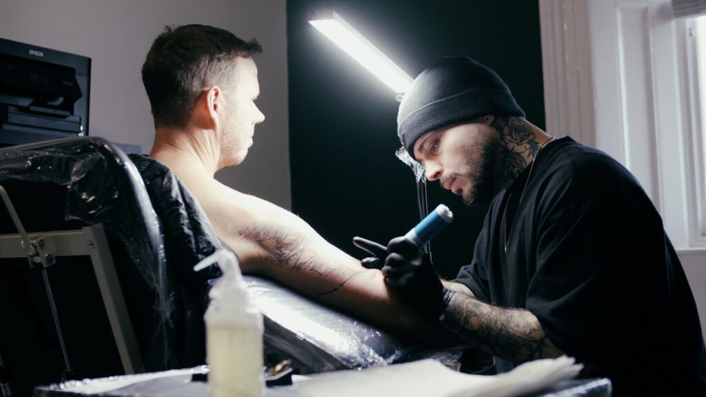 Image is of a male client getting a tattoo on his upper arm by a male tattoo artist wearing all black. The client is getting his first tattoo.