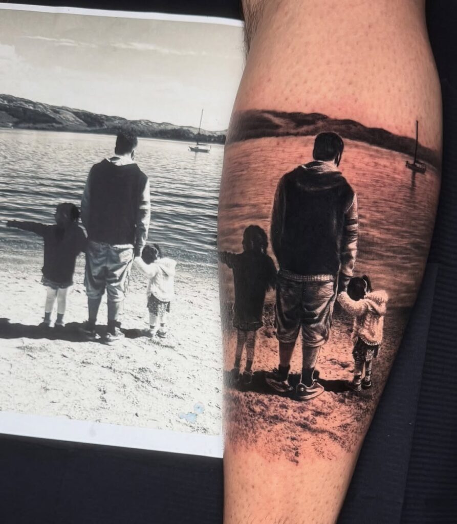 A black and gray leg tattoo of a man holding hands with his two children, standing on a beach, looking out to the sea.