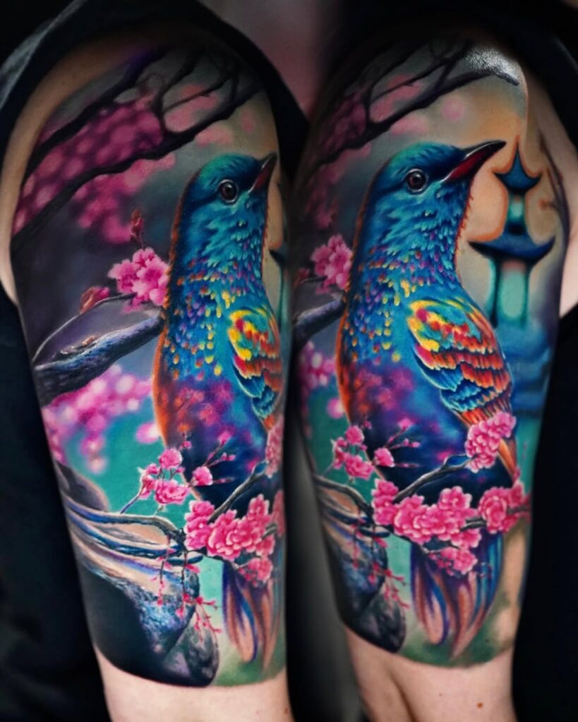 A colourful blue bird tattoo in front of pink cherry blossom by Joan Pereira.