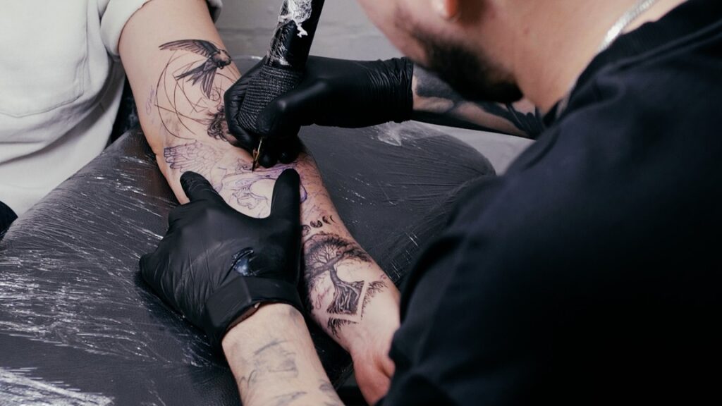 A male tattoo artist tattooing a clients forearm. It shows a swallow and a tree of life in black and gray. 