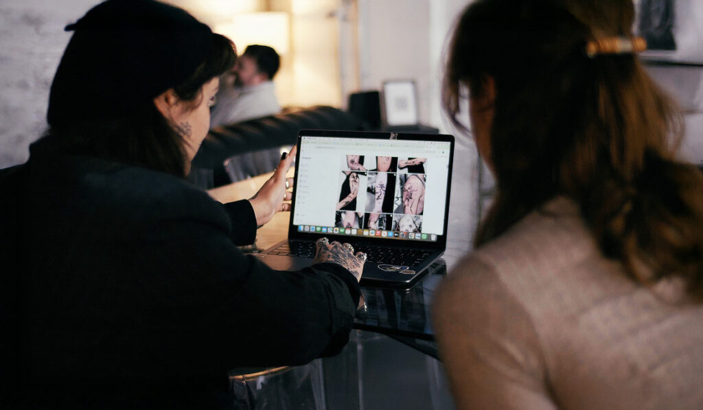 A female tattoo consultant and female client look at a laptop where a tattoo artist's portfolio is on screen.