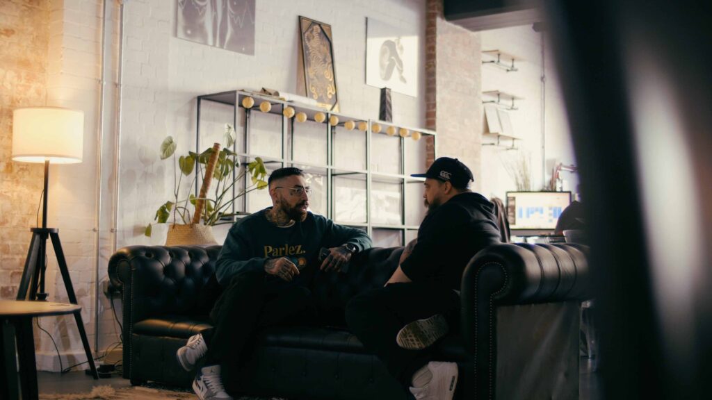 A male tattoo consultant sits opposite a male client on a sofa having a tattoo consultation. 