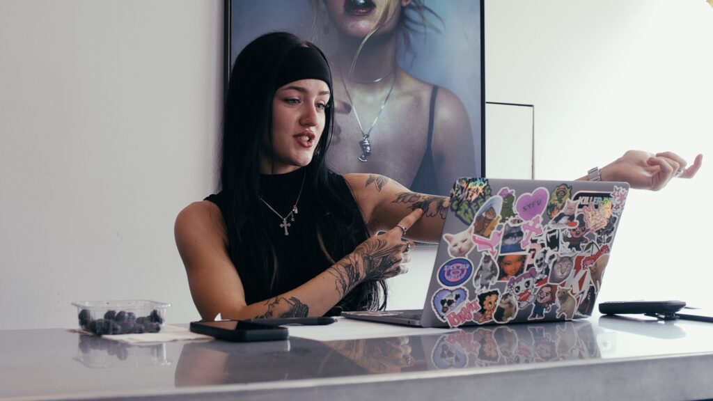 A female tattoo consultant is looking at a laptop during a video tattoo consultation. She is pointing to her arm. 