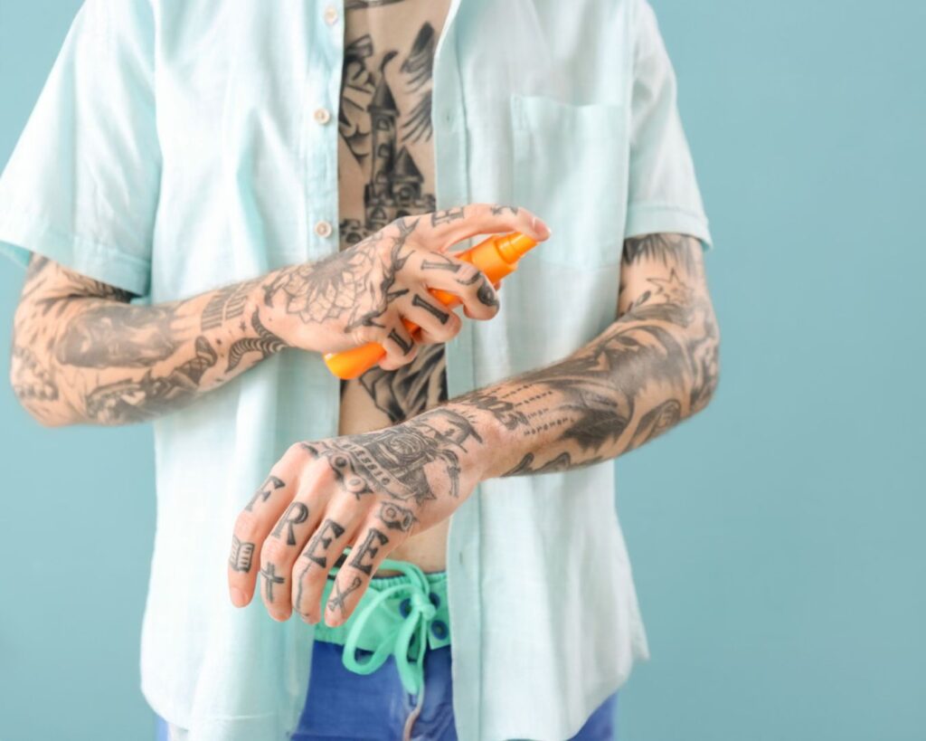 A tattooed male sprays sunscreen onto his tattooed arm.