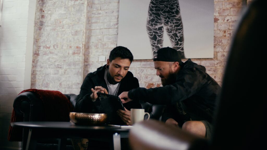 A male tattoo artist and client sit together on a sofa looking at an iPad during a tattoo consultation. 