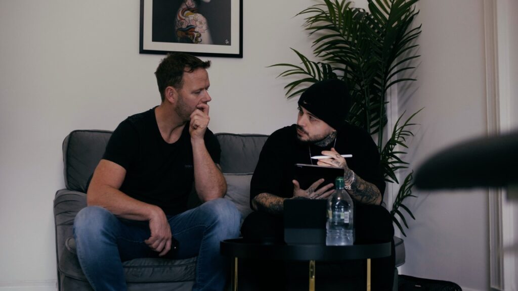 A male client and tattoo artist sitting on a sofa during a tattoo consultation. The artist is holding an iPad and pen and looking at the client. 