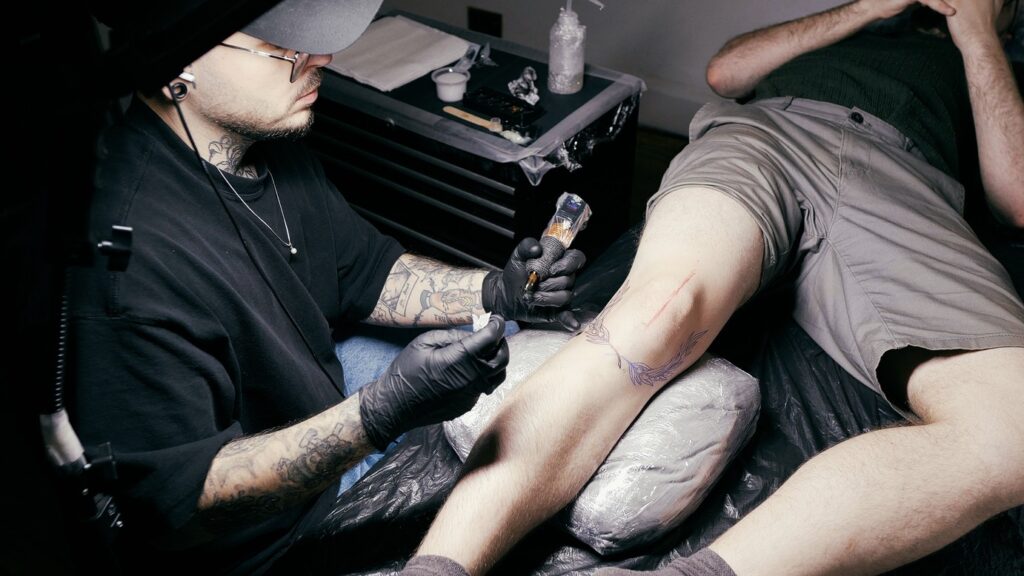 A male artist tattoos the knee of a client. 